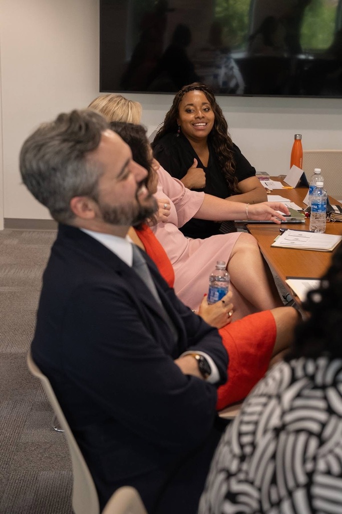 A close-up view of meeting participants seated at conference tables. A woman in a black top smiles in the background while a man in a navy suit with an orange tie is shown in the foreground, both engaged in discussion.