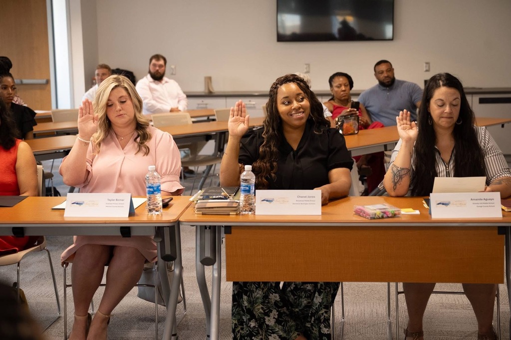 Three women raise their hands while seated at a conference table with name placards and water bottles. The woman in the center, wearing a black top, smiles as she participates in what appears to be a group activity or vote.