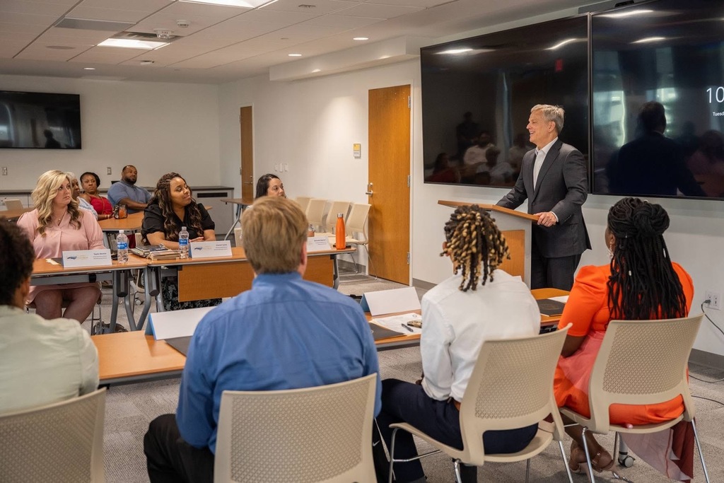 A man in a dark suit presents to a diverse group of educators seated at tables in a modern conference room. Participants have name placards, water bottles, and materials in front of them. A large screen displays "10" in the background.