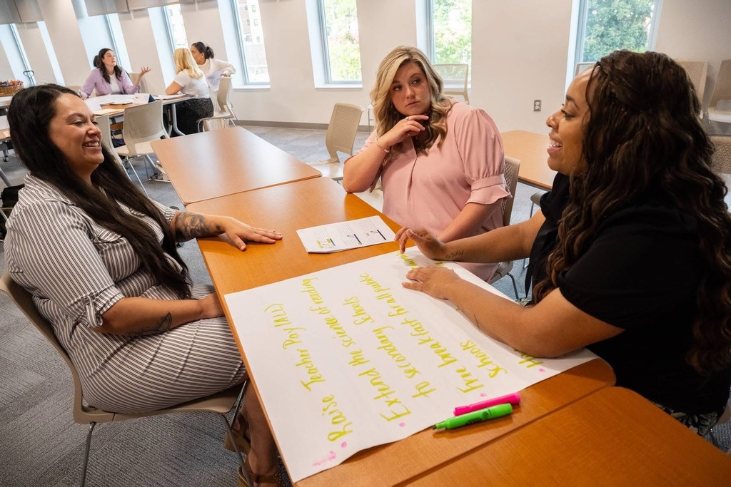 Three women collaborate around a large sheet of paper with yellow sticky notes arranged on it. They appear to be engaged in a brainstorming or planning activity, with colored markers visible on the table.