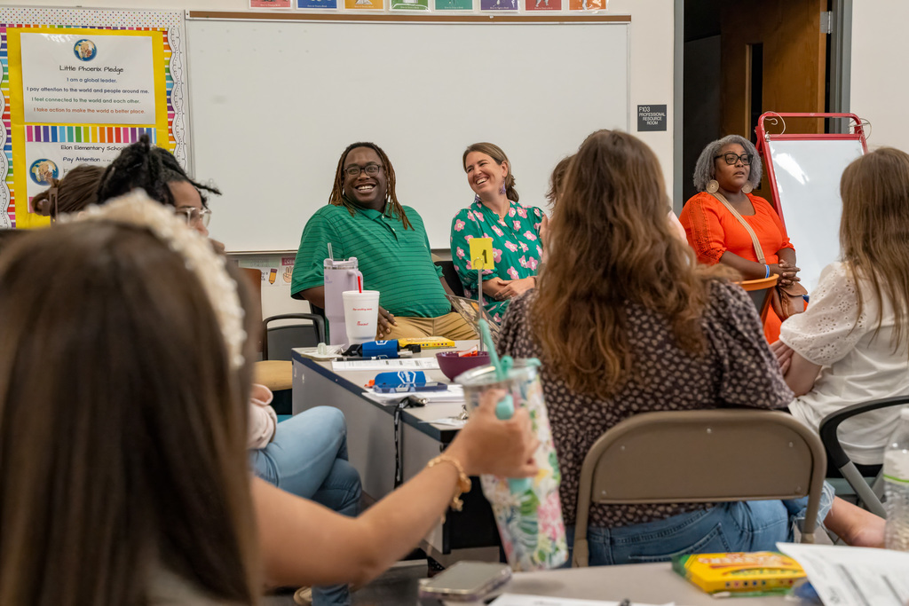 Beginning teachers sit in a classroom during orientation, smiling and engaged as presenters lead a session. Classroom walls display educational posters and the "Little Phoenix Pledge" is visible in the background.