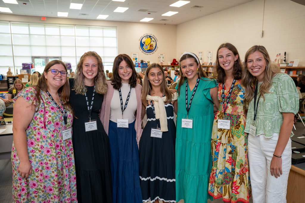 A large group of seven beginning teachers pose together in a school library, all wearing orientation lanyards and displaying bright smiles. The school's logo is visible on the wall behind them.