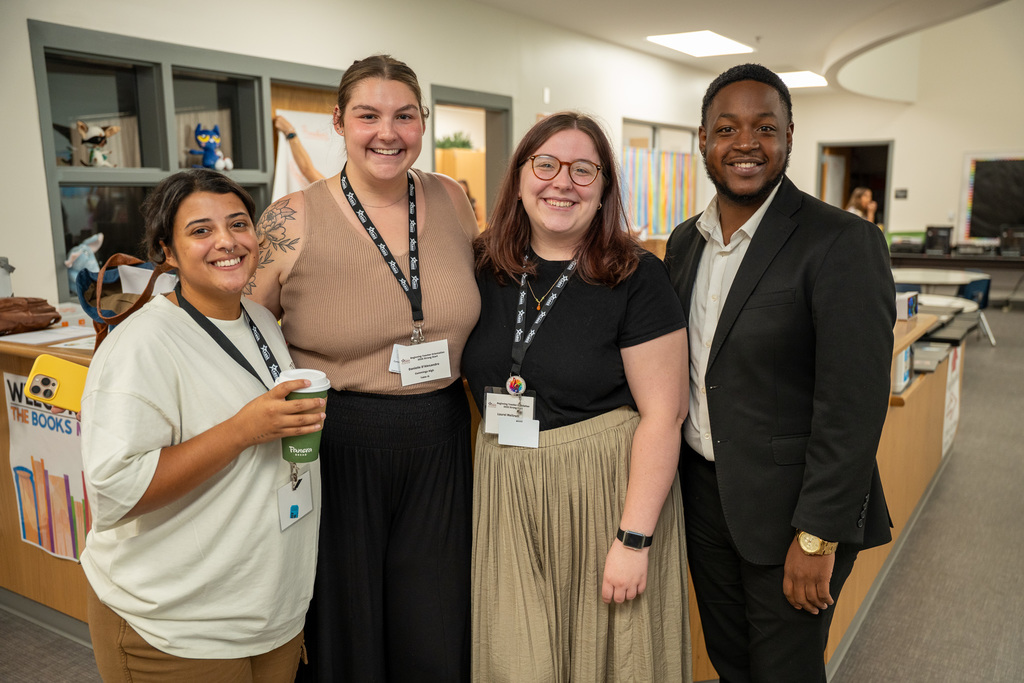 Four new beginning teachers pose together in a school library, wearing name badges and lanyards. They stand smiling among book displays and educational materials.