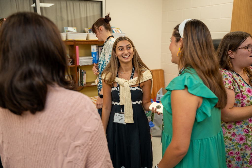 Beginning teachers engage in conversation during a networking portion of the orientation, standing in a classroom with educational materials and bookshelves visible in the background.