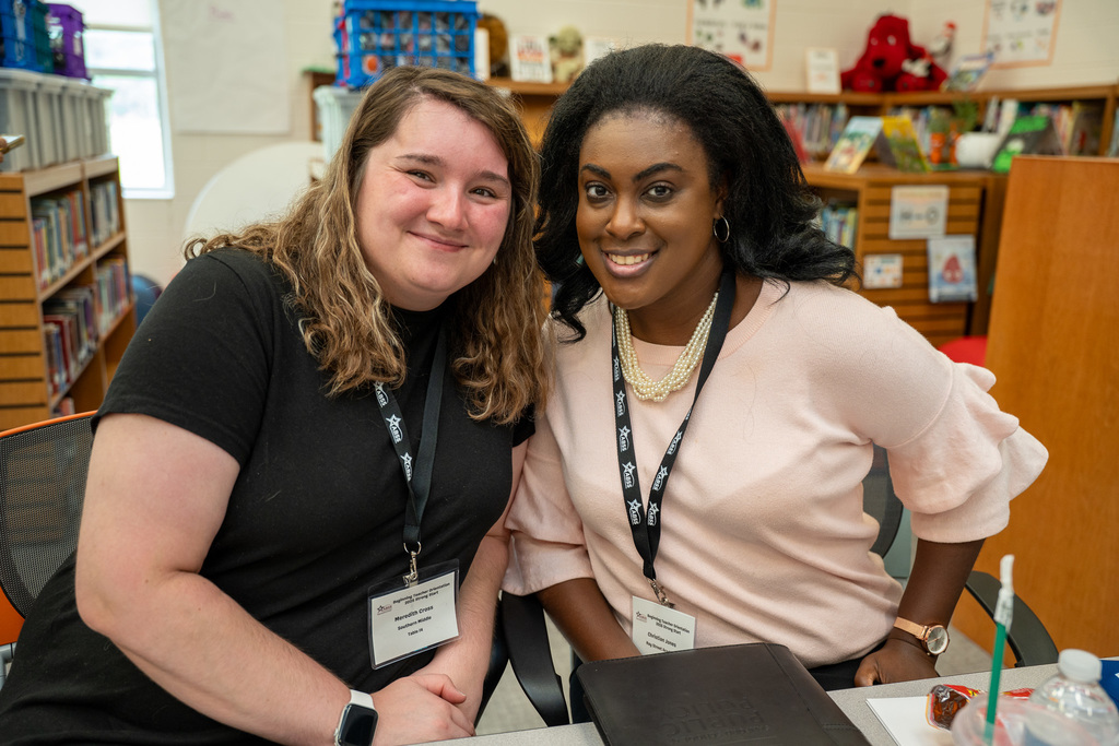 Two beginning teachers pose together in a school library, sitting at a table with educational materials nearby. Both are wearing orientation name badges and appear relaxed and happy.