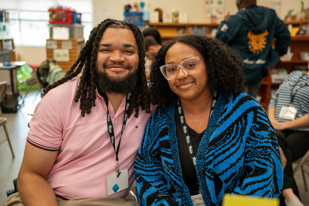 Two beginning teachers sit together at a table in a classroom setting, both wearing orientation lanyards and smiling at the camera during a break in the day's activities.