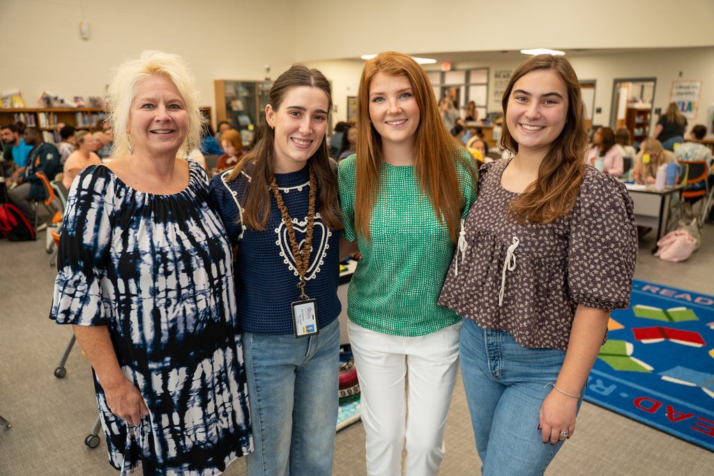 A group of four people, including beginning teachers and what appears to be a mentor or coordinator, stand together in a busy school cafeteria during the orientation lunch period.