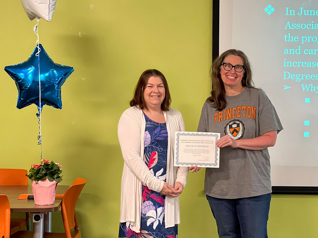 Math teacher Jesica Fitzgerald holding an award certificate in front of a yellow classroom backdrop. Another educator stands beside her.