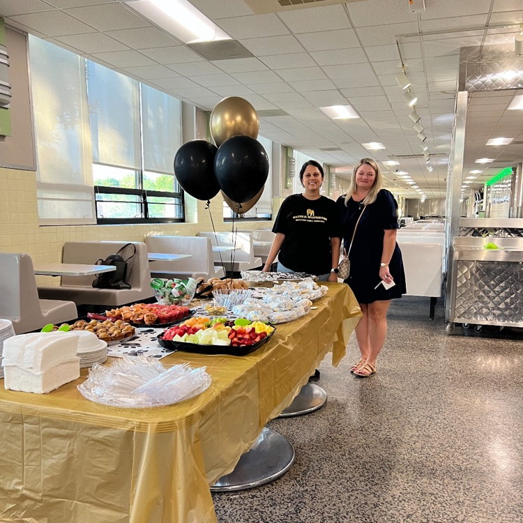 Breakfast foods on a gold tablecloth with two ladies standing at the end of the table