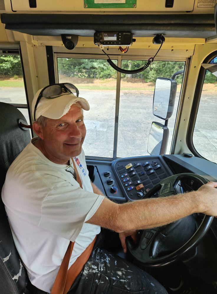 Bus driver in a white t-shirt and hat sitting in the driver's seat with one hand on the wheel.  The bus is parked.