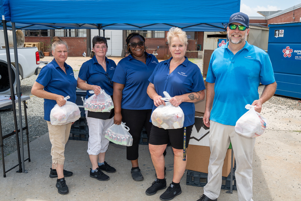Five school nutrition workers in blue shirts hold bags of food for families at a summer food program serving site