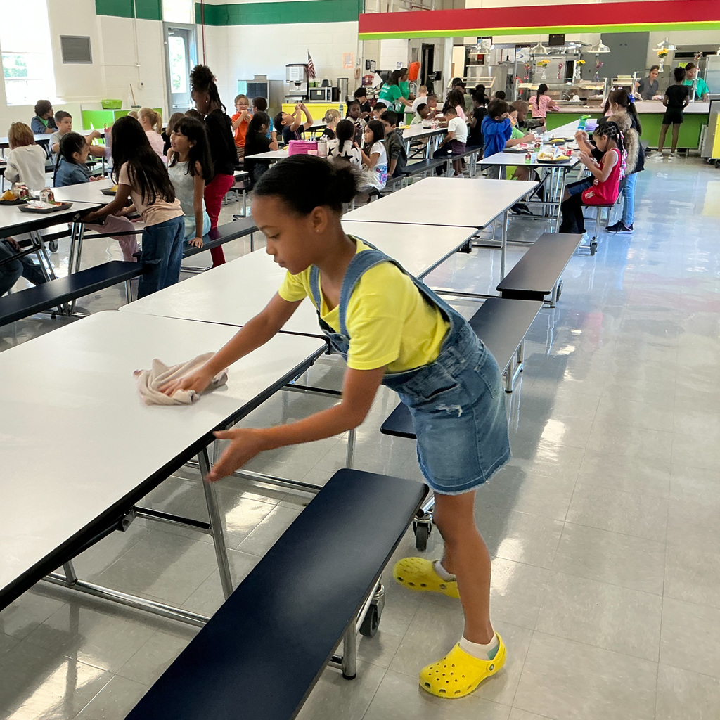 A young girl in a yellow shirt and denim overalls wipes down a cafeteria table