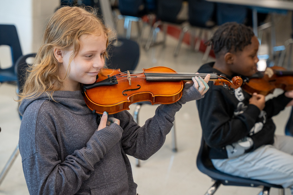 A blonde elementary student in a gray hoodie holding a violin in playing position, with other students visible in the background of a music classroom.