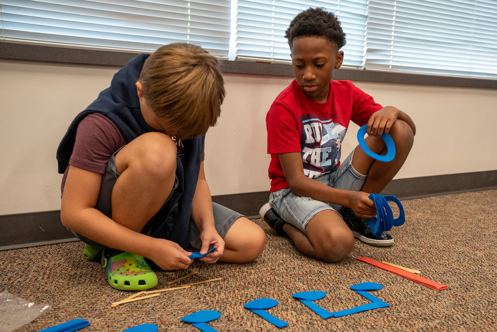 Two elementary boys sitting on a classroom floor working with blue musical note shapes and other colorful educational materials spread around them.