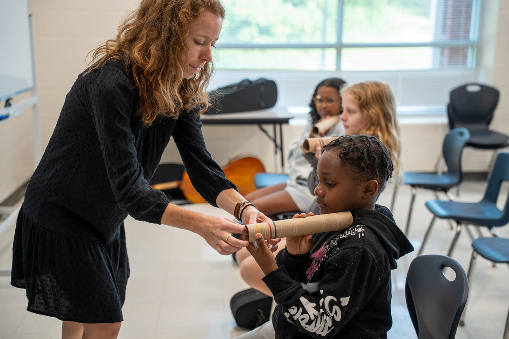A female teacher with curly hair helping an elementary student holding a paper tube, with other students visible in the background.