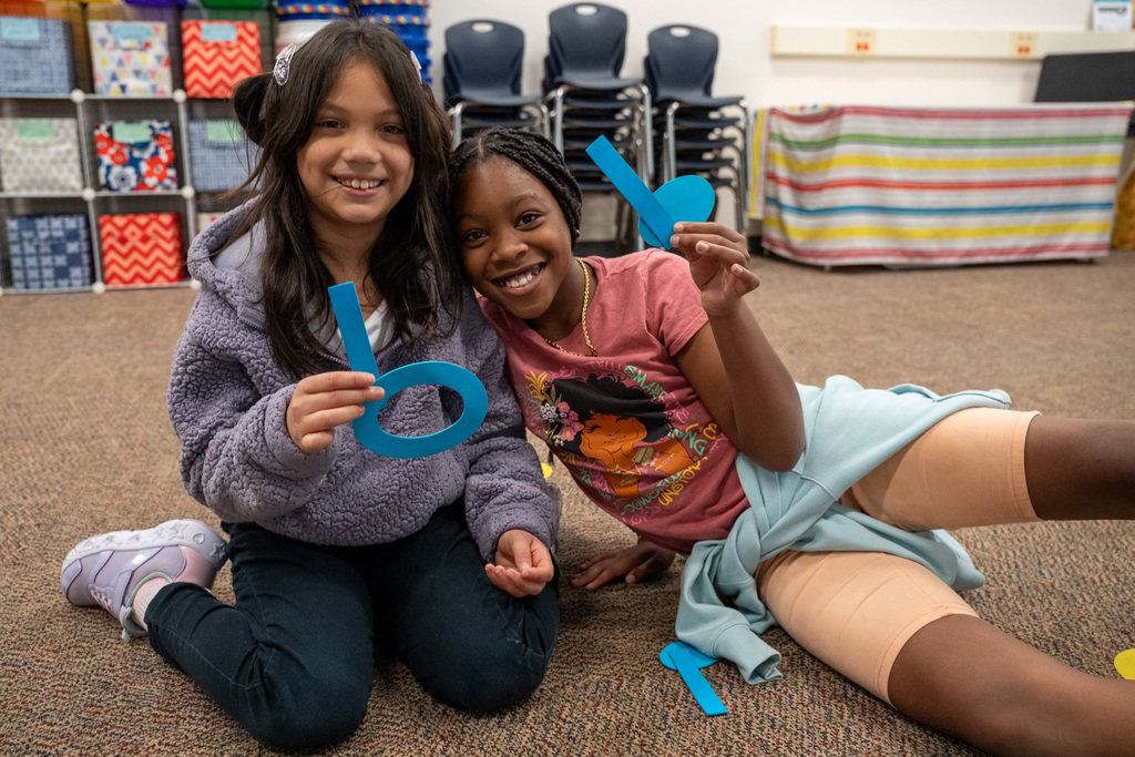 Two elementary students sitting cross-legged on a classroom carpet, smiling at the camera while holding blue musical note cutouts. Classroom storage bins and chairs are visible in the background.
