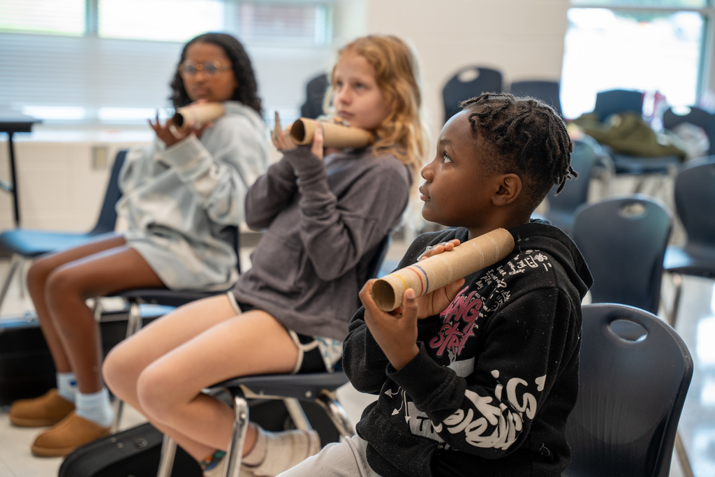 Three elementary students sitting in chairs, each holding cardboard paper towel tubes, appearing to be participating in a music lesson.