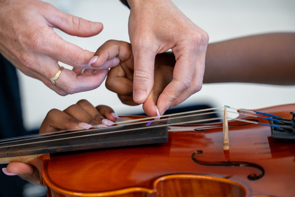 Close-up of adult hands guiding a child's small hands on violin strings, demonstrating proper finger placement technique on the instrument's fingerboard.