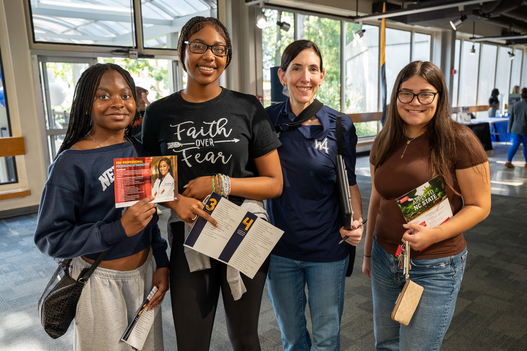 Four people pose together in a modern college building interior - three high school students and one adult. The students are holding various college brochures and informational materials, with large windows and contemporary architecture visible behind them.