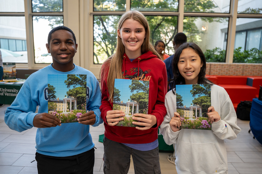 Three high school students pose together indoors, each holding identical college brochures featuring a campus building with columns. The students are wearing casual clothing including hoodies and sweatshirts.