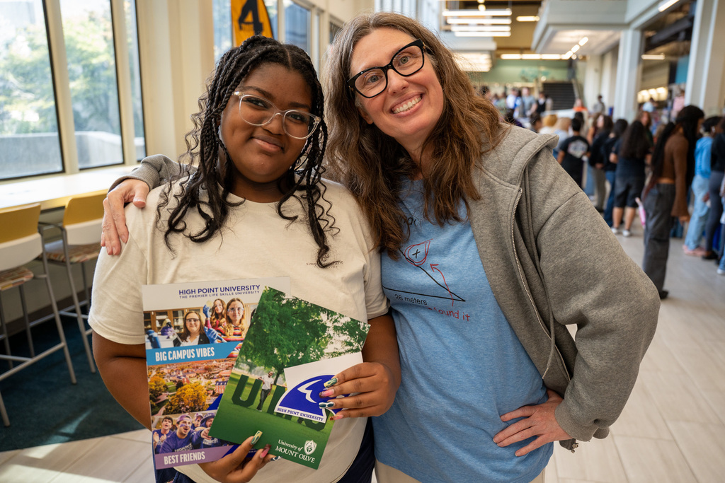 A high school student and adult advisor pose together in a school hallway, both smiling. The student holds college brochures including materials from High Point University and University of Mount Olive. Other students can be seen in the background.