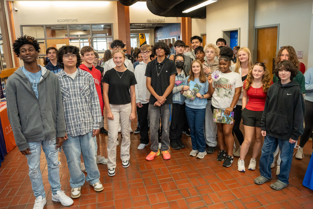 A large group of high school students gathers in what appears to be a college bookstore or student center, with "BOOKSTORE" signage visible in the background. The diverse group is smiling and some are holding college materials.