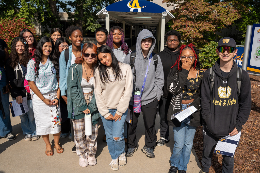 A large group of diverse high school students stands together outside Alamance Community College, with the college's blue and yellow signage visible behind them. Students are holding college fair materials and information packets.
