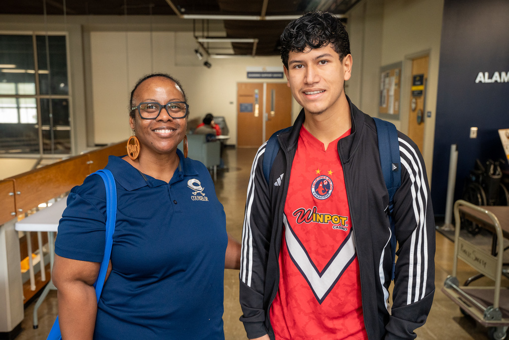 An adult counselor or advisor poses with a high school student. The counselor wears a blue polo shirt with college branding, while the student wears a red sports jersey with "Winpot" visible on it.