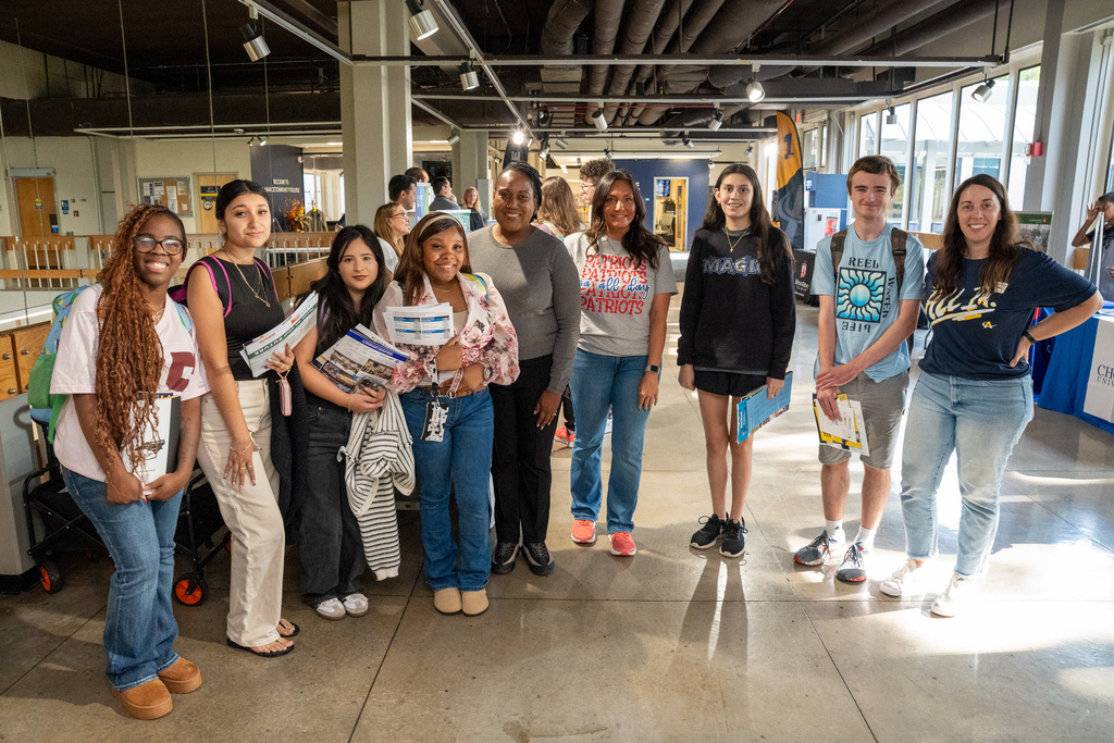 A diverse group of high school students poses with an adult advisor in a modern college building. Students are holding various college brochures and materials, with the contemporary interior architecture of the building visible in the background.
