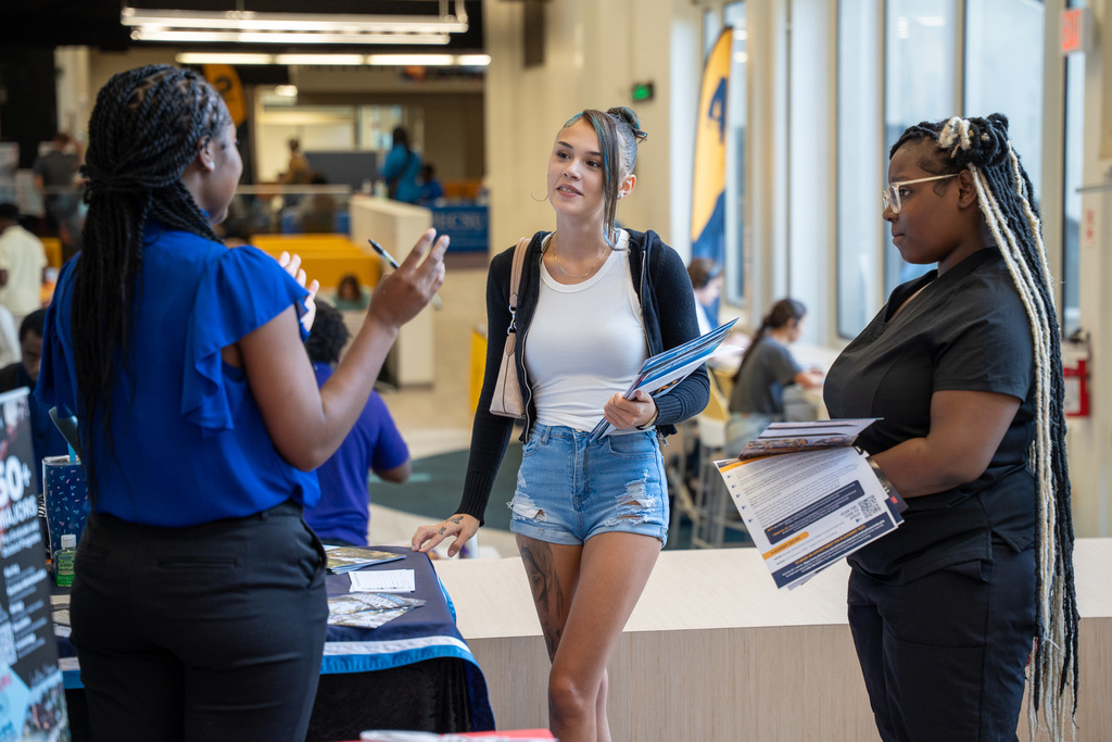 Two high school students engage in conversation with a college representative at an information table. The representative, wearing blue, appears to be explaining programs while students hold informational materials.