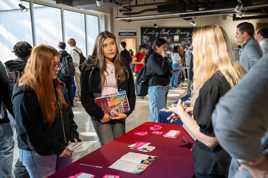 High school students interact with college representatives at information tables in a busy college fair setting. Students are examining materials while representatives provide information about their institutions.