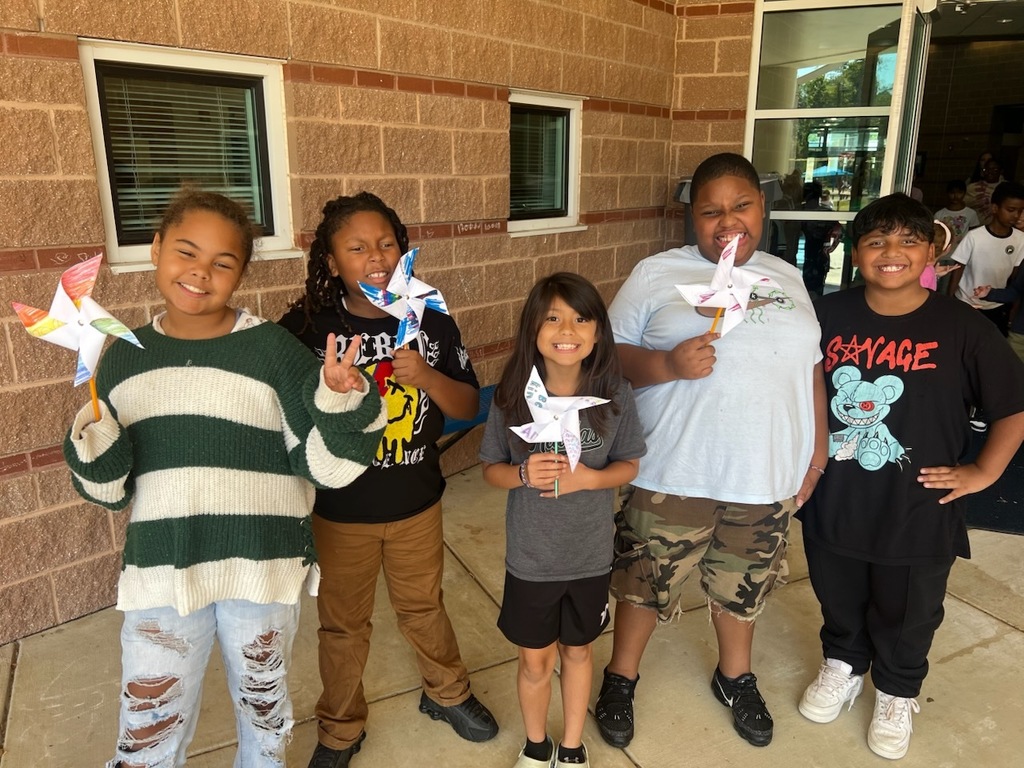 Five smiling elementary students stand outside a brick school building, each proudly holding their colorful paper peace pinwheels they made for International Peace Day. The students are wearing casual clothes and display their handcrafted pinwheels on wooden sticks.
