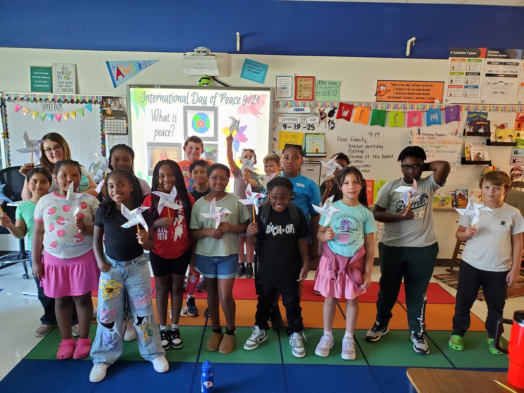 A diverse group of 4th grade students at Highland Elementary stand together in their colorful classroom holding handmade peace pinwheels. Behind them is a whiteboard display about International Day of Peace 9/21 with the question 'What is peace?' The classroom walls are decorated with educational posters and student work.