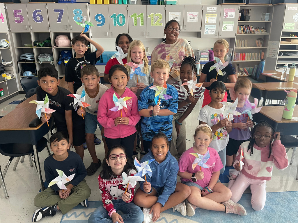 A classroom full of 4th grade students pose with their teacher, all holding peace pinwheels they created. The organized classroom features numbered storage cubbies (5-12 visible) along the wall and educational materials. Students are arranged in multiple rows, sitting and standing, proudly displaying their colorful paper pinwheel creations.