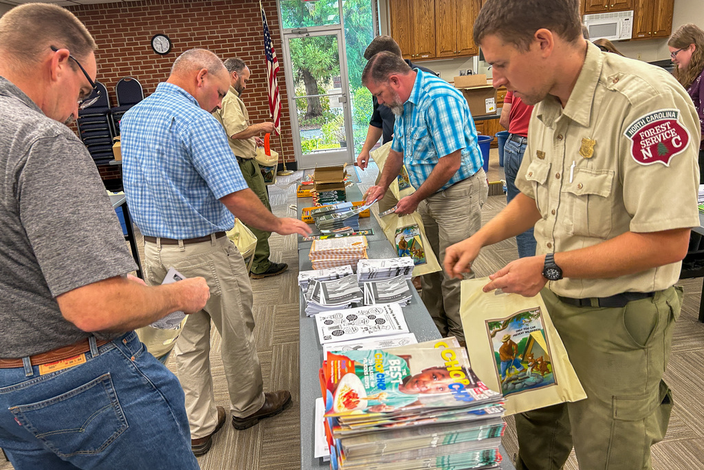 Several men in a community center kitchen area sorting through educational materials on tables. One man wears a North Carolina Forest Service uniform. They're organizing various publications, books, and educational resources spread across multiple tables.