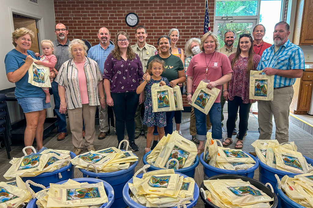 A large group of volunteers and officials pose together in a community center room with brick walls. They're holding canvas tote bags with colorful agricultural-themed designs. In front of them are multiple blue plastic bins filled with hundreds of similar tote bags, ready for distribution.