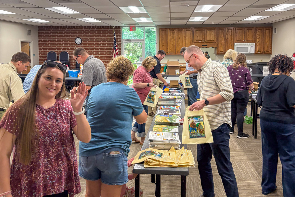 A busy scene in a community center with volunteers packing educational materials into tote bags. Multiple people work at tables covered with agriculture-themed publications and materials. A young woman in a floral dress waves at the camera from the left side.