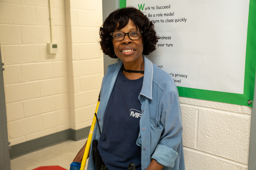 Smiling custodian wearing blue work shirt over MFM uniform standing in a school hallway next to a motivational poster about kindness and success