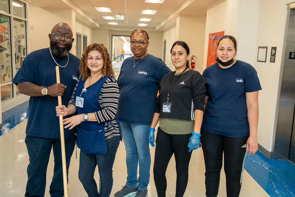 Five custodial staff members posing together in a school hallway, wearing MFM uniforms and holding cleaning equipment including a broom