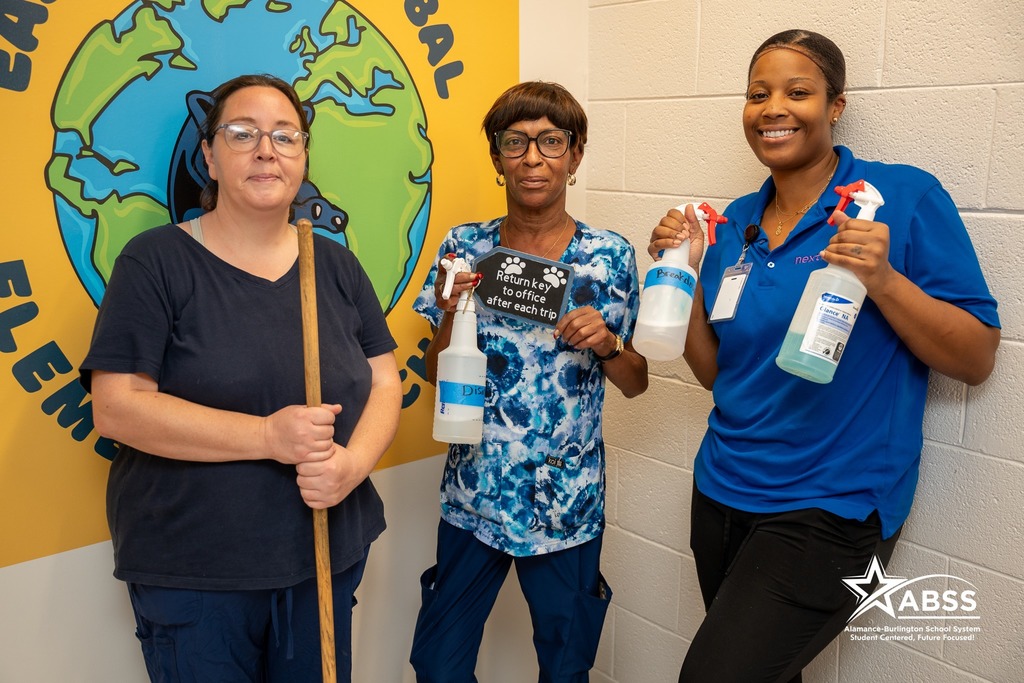Three custodial staff members posing in front of a colorful globe mural, holding cleaning supplies including a broom and spray bottles, with one staff member holding a sign that reads "Return key to office after each trip"Retry