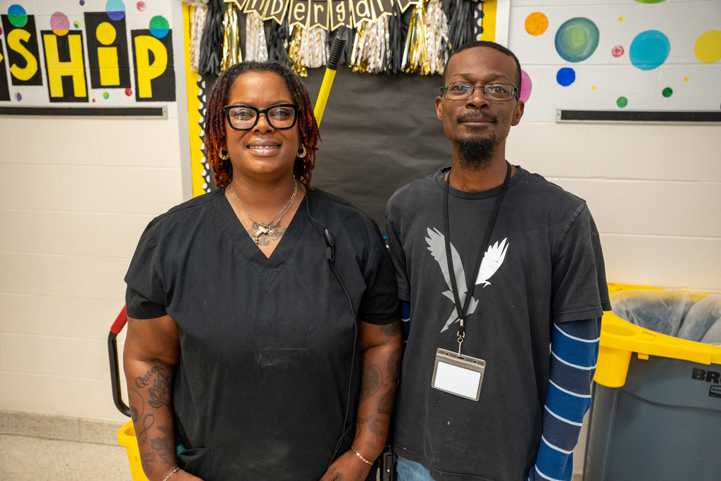 Two custodians smiling together in front of a colorful bulletin board decorated with "SHIP" lettering and celebration streamers