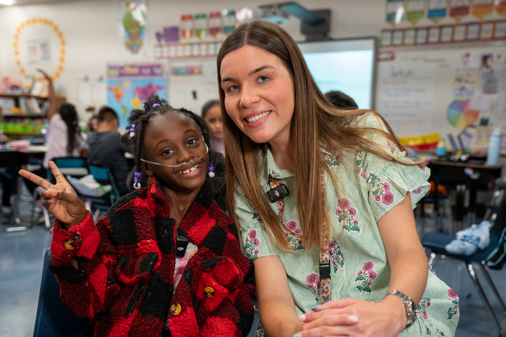 Ms. Steinki kneels beside a young student who is making a peace sign with her hand. Both are smiling at the camera. The student wears a red and black plaid jacket, and the classroom activity continues behind them.