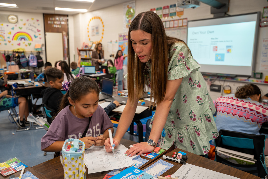 Ms. Steinki leans over a student's desk, providing one-on-one instruction as the student works on a math worksheet. The teacher is pointing to the student's work while other classmates work independently at their desks in the background. A smartboard displaying a math lesson is visible on the wall.