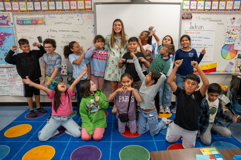 Image 1: Teacher Anna Sophia Steinki stands at the front of her classroom surrounded by enthusiastic elementary students striking celebratory poses. The students are arranged in two rows on a colorful circular carpet, with some kneeling and others standing, all making excited gestures with their hands raised. A whiteboard behind them shows a division equation (2,800 ÷ 4 = 700) and the date October 1, 2025.