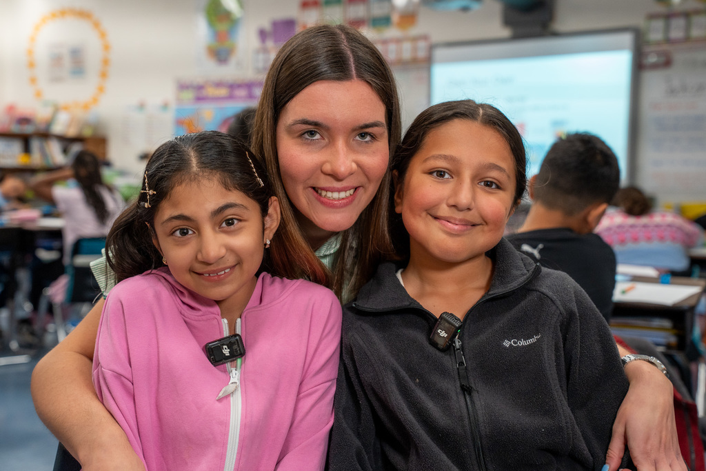 Ms. Steinki poses with two smiling students who have their arms around her. All three are looking at the camera with warm smiles in the classroom setting with other students visible working in the background.