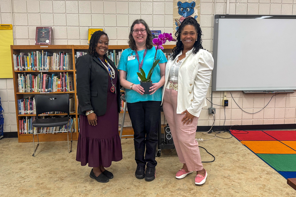 An educator of excellence holding a colorful purple flower arrangement surrounded by ABSS staff in a school library setting