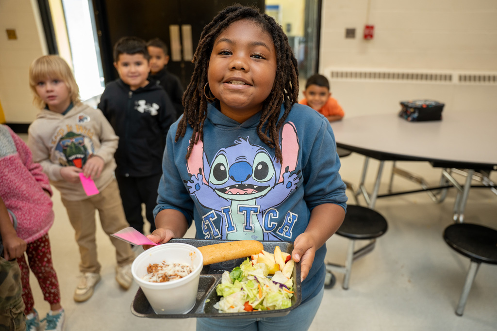 Student in Stitch sweatshirt carrying lunch tray with spaghetti, breadstick, and salad through cafeteria line