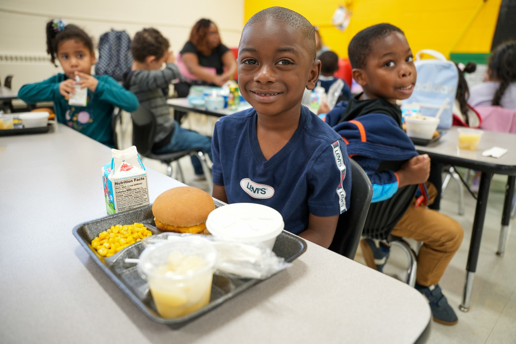 Student smiling at lunch table with tray containing chicken sandwich, corn, milk, and fruit cup in elementary school cafeteria