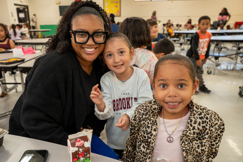 School staff member in glasses posing with two young students in elementary school cafeteria during lunch period