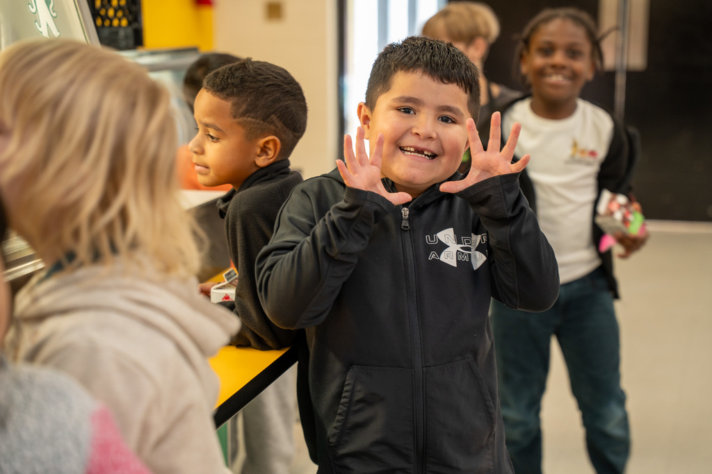 Student in black Under Armour jacket waving both hands at camera with big smile while standing in cafeteria